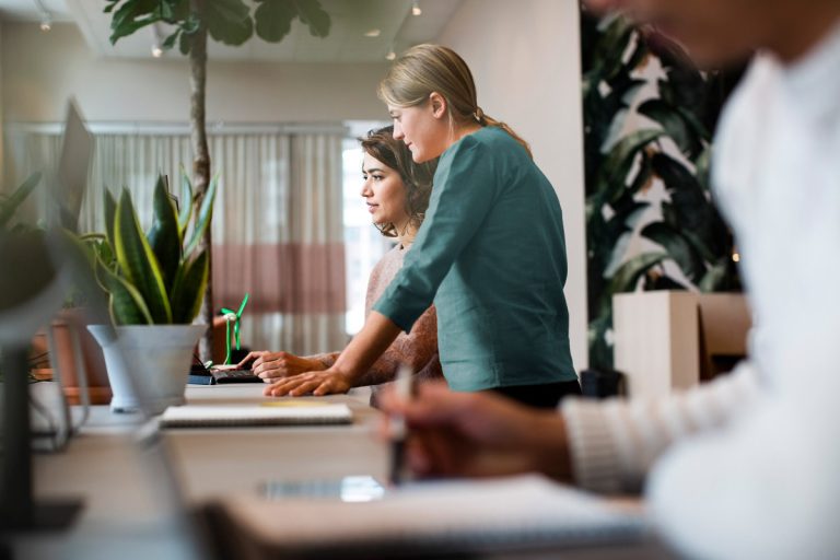 Deux collaboratrices, l’une debout et l’autre assise, travaillant ensemble sur un ordinateur dans un bureau moderne, un troisième collègue flou au premier plan